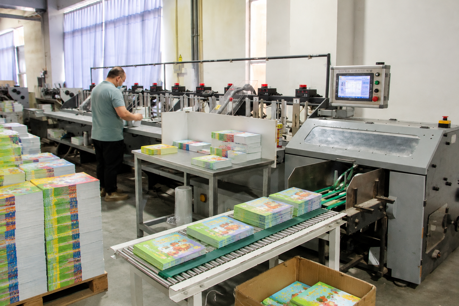 Children's book printing factory with workers operating book finishing equipment and colorful children's books moving along a conveyor line