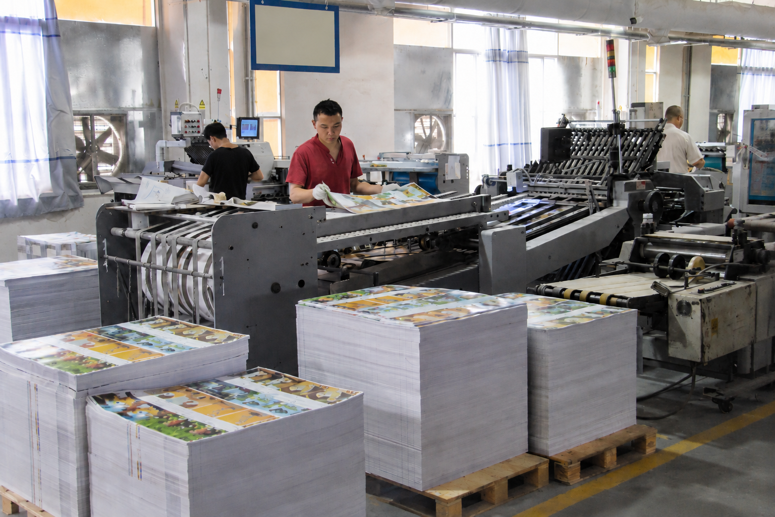 Workers inspecting printed children's book sheets on an industrial folding and finishing line at a book printing factory